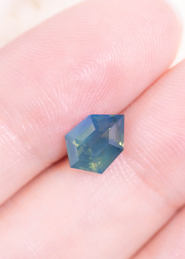 Loose opalescent greenish teal blue sapphire resting on a fingertip for scale, showing the shield step cut and the teal-blue body color with soft, opalescent depth.
