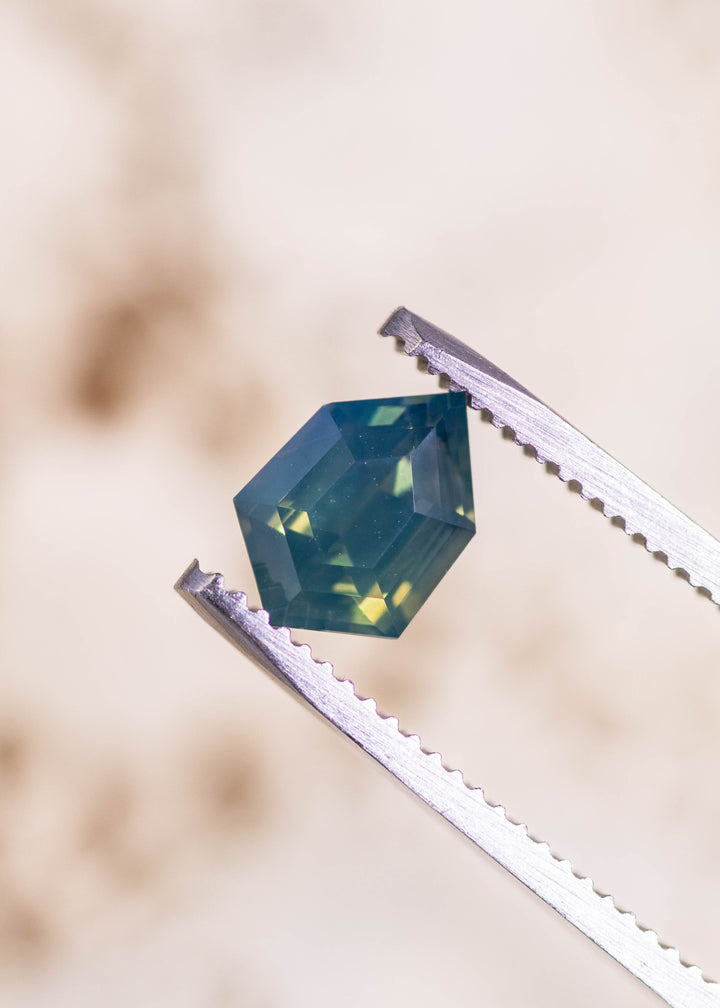 Loose opalescent greenish teal blue sapphire from Diego, Madagascar held in tweezers, showing the geometric shield step cut with crisp corners and bright green flashes.