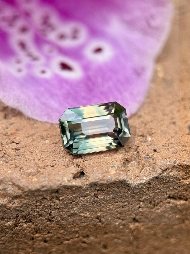 Close-up of a 1.60 carat sapphire in an emerald cut, capturing its earthy parti colors under lighting—set against a slate gray stone slab.