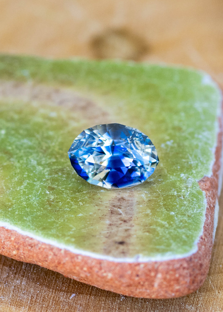 1.60ct blue and white bi color parti sapphire from Sri Lanka resting on a green piece of sea pottery.