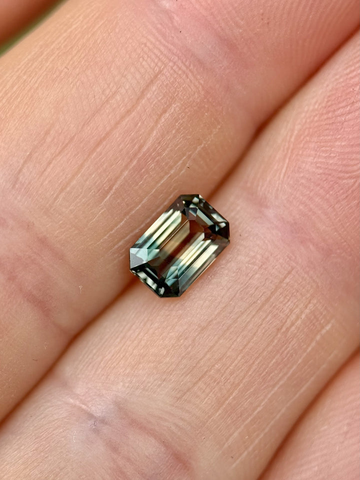 1.60 carat untreated sapphire from Sakaraha, Madagascar, showing a fluorescent color change from earthy peach to lagoon green, held between fingers in sunlight with green foliage in the blurred background.