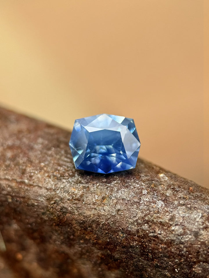 1.60-carat sky-denim and royal-blue opalescent parti sapphire from Diego, Madagascar displayed on textured bark, showing its precision custom geometric cushion cut and natural untreated color zoning.