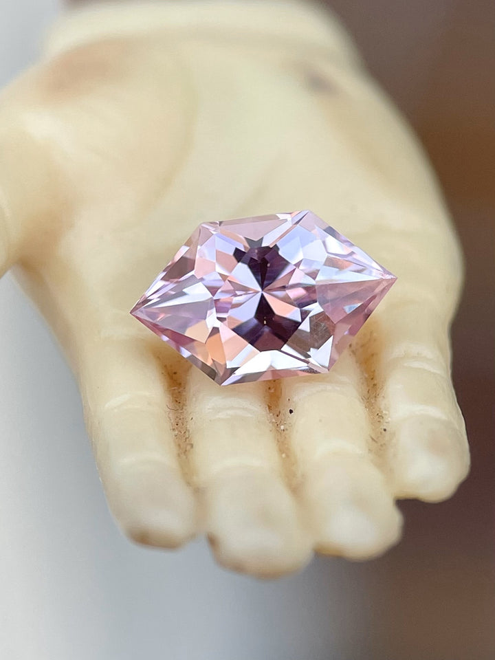Close-up view of a 1.61 carat blush pink and peach sapphire, featuring a modified brilliant hexagon cut, resting on a vintage hand sculpture for a refined presentation. The gemstone displays a delicate blend of blush pink and peach tones.