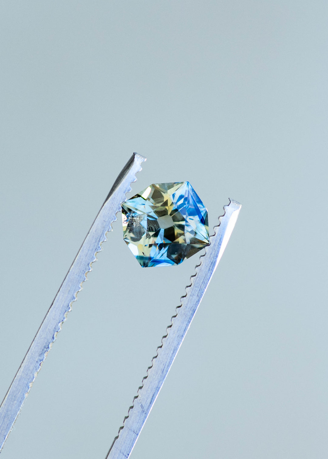 1.62ct teal yellow parti custom old mine cushion cut sapphire held in gem tweezers showing off the open culet and parti color zoning.
