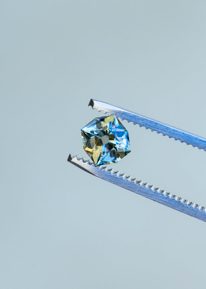 1.62ct teal yellow parti custom old mine cushion cut sapphire held in gem tweezers showing off teal and yellow parti color zoning.