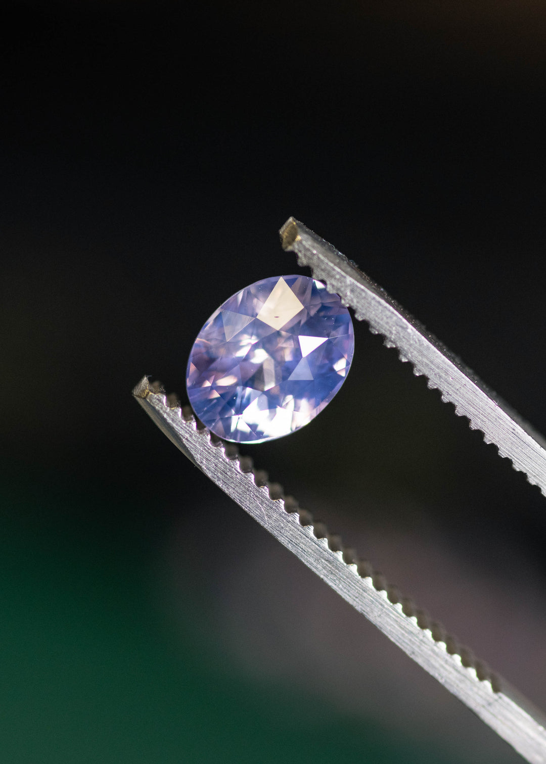 Loose pinkish purple sapphire from Ratnapura held in tweezers, showing reflective mix-cut facets and soft violet-rose color shift.