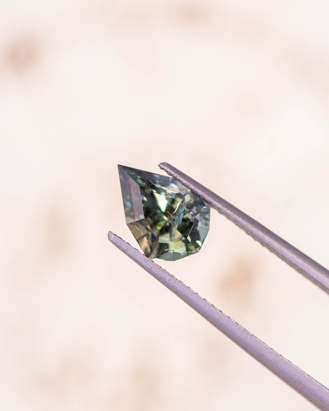 1.64 carat teal green yellow tanzanite held delicately in gemstone tweezers against a soft neutral background, showing detailed precision faceting and bright reflections in this untreated geometric mixed cut shield from Merelani Hills, Tanzania.