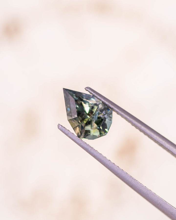 1.64 carat teal green yellow tanzanite held delicately in gemstone tweezers against a soft neutral background, showing detailed precision faceting and bright reflections in this untreated geometric mixed cut shield from Merelani Hills, Tanzania.