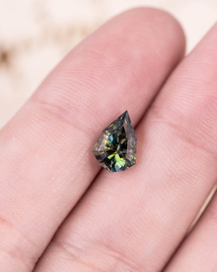 1.64 carat tanzanite resting on fingertip, displaying vivid yellow, green, and teal hues with intricate custom geometric mixed cut shield facets, untreated gemstone from Merelani Hills, Arusha Region, Tanzania.