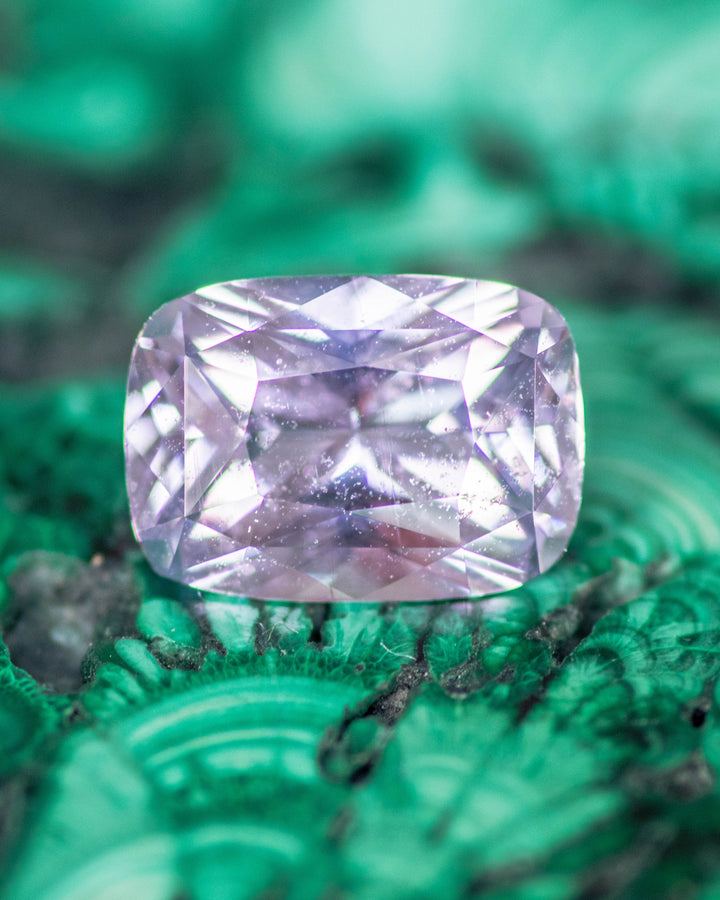 1.65 carat violet, purple, and pink sapphire resting on malachite background, highlighting its vibrant cushion brilliant cut facets and internal reflections, untreated gemstone from Sakaraha, Madagascar.
