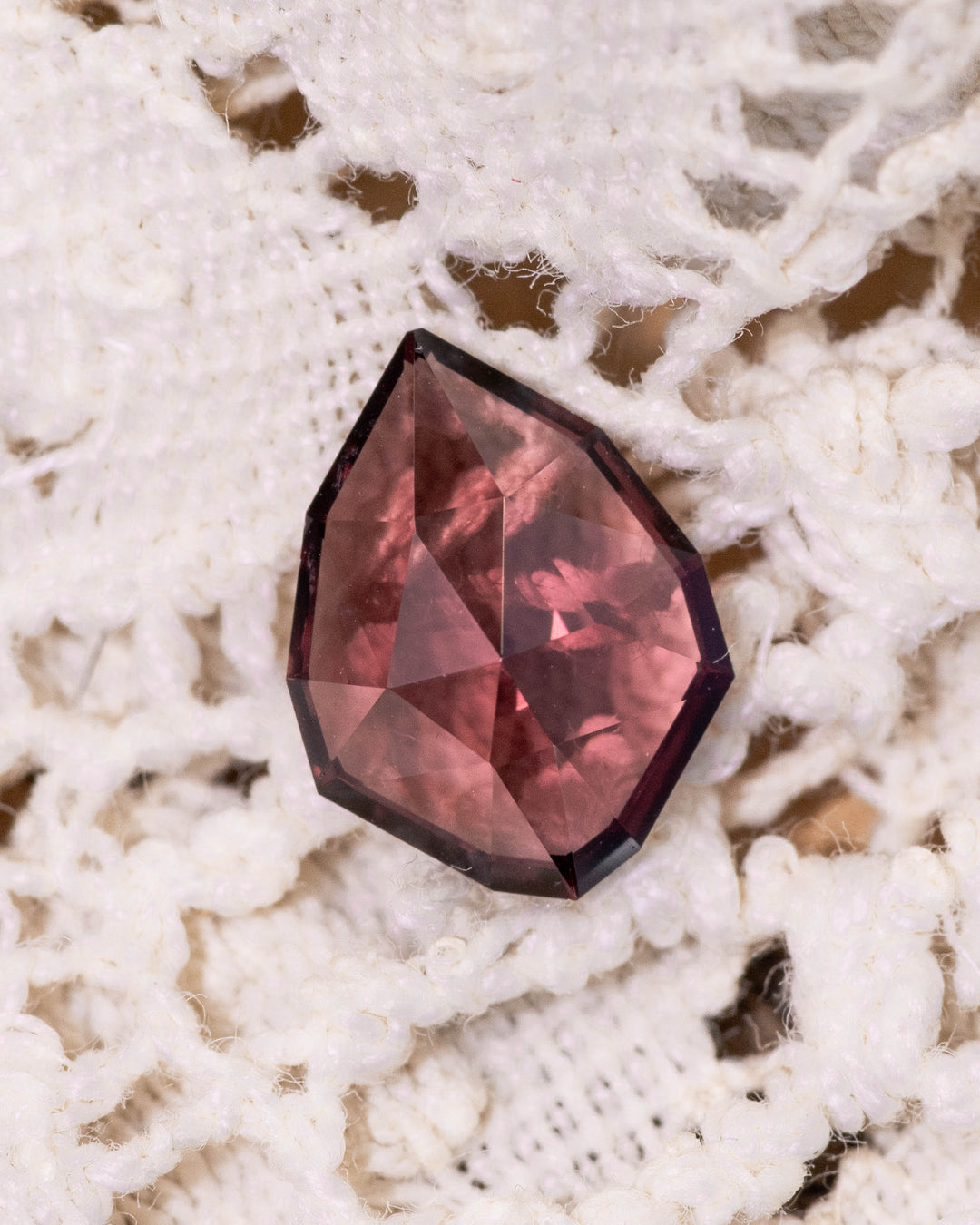 1.66 carat red and orange garnet resting on white lace fabric, showing the vivid hues and facets of the custom geometric rose cut shield, untreated garnet from Kenya