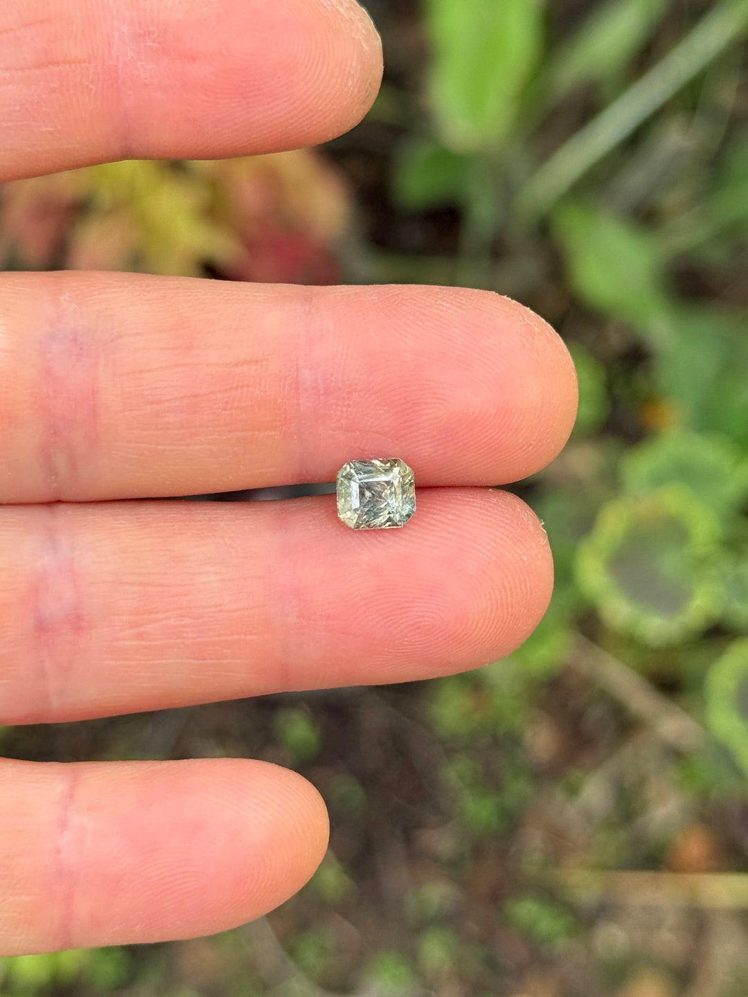 1.67 carat color-change sapphire balanced on fingertips for size reference, revealing its custom squared emerald cut and subtle shift from earthy green to red under daylight, natural and untreated gem from Umba River, Tanzania.