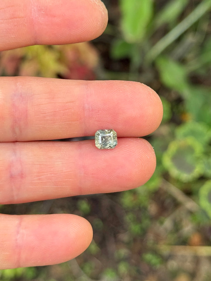 1.67 carat color-change sapphire balanced on fingertips for size reference, revealing its custom squared emerald cut and subtle shift from earthy green to red under daylight, natural and untreated gem from Umba River, Tanzania.