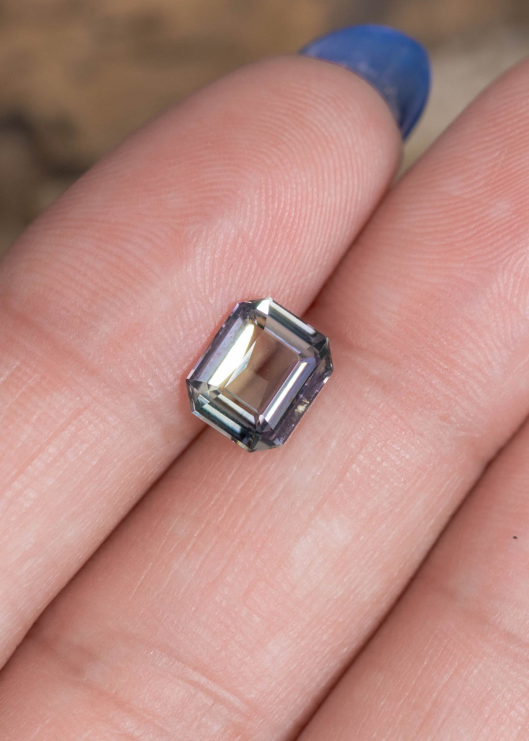 1.67 carat teal blue green purple Tanzanite custom modified emerald cut resting on finger, Tanzania origin gemstone highlighting elongated rectangular shape, mirror-like step faceting and vibrant trichroic brilliance for bespoke jewelry