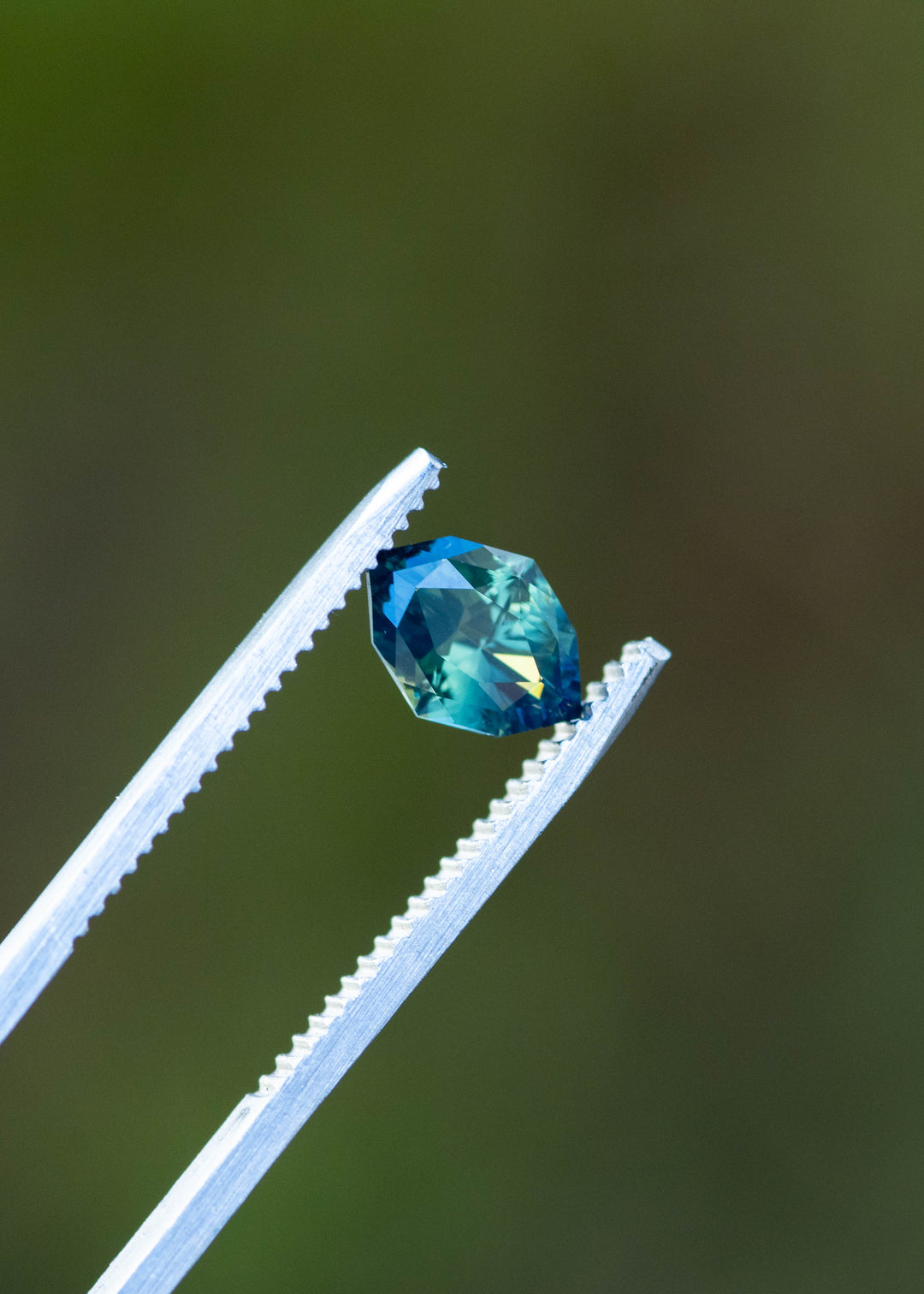 1.68ct green teal geometric marquise sapphire from Diego, Madagascar held in gem tweezers against green background under natural lighting.