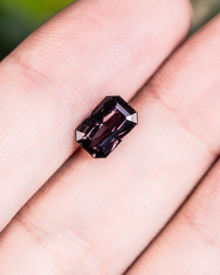 1.70 carat violet brown sapphire resting on a fingertip under natural daylight, revealing its rectangular emerald cut facets and natural clarity, untreated gemstone from Umba River Valley, Tanzania.