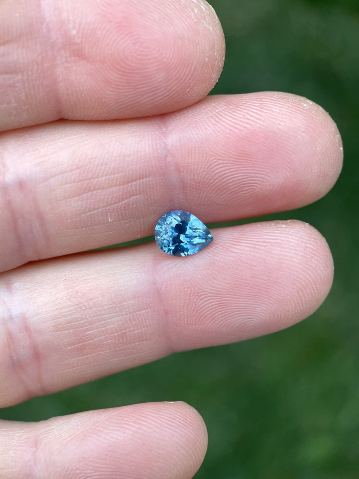 1.70 carat blue sapphire displayed on fingertip in natural light, showcasing its custom brilliant pear cut and vivid blue hue.