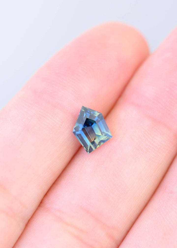 1.70 carat Madagascar parti sapphire resting on fingertips for scale, showing its unconventional freeform outline, broad step facets and natural blue, teal, green and yellow coloration.