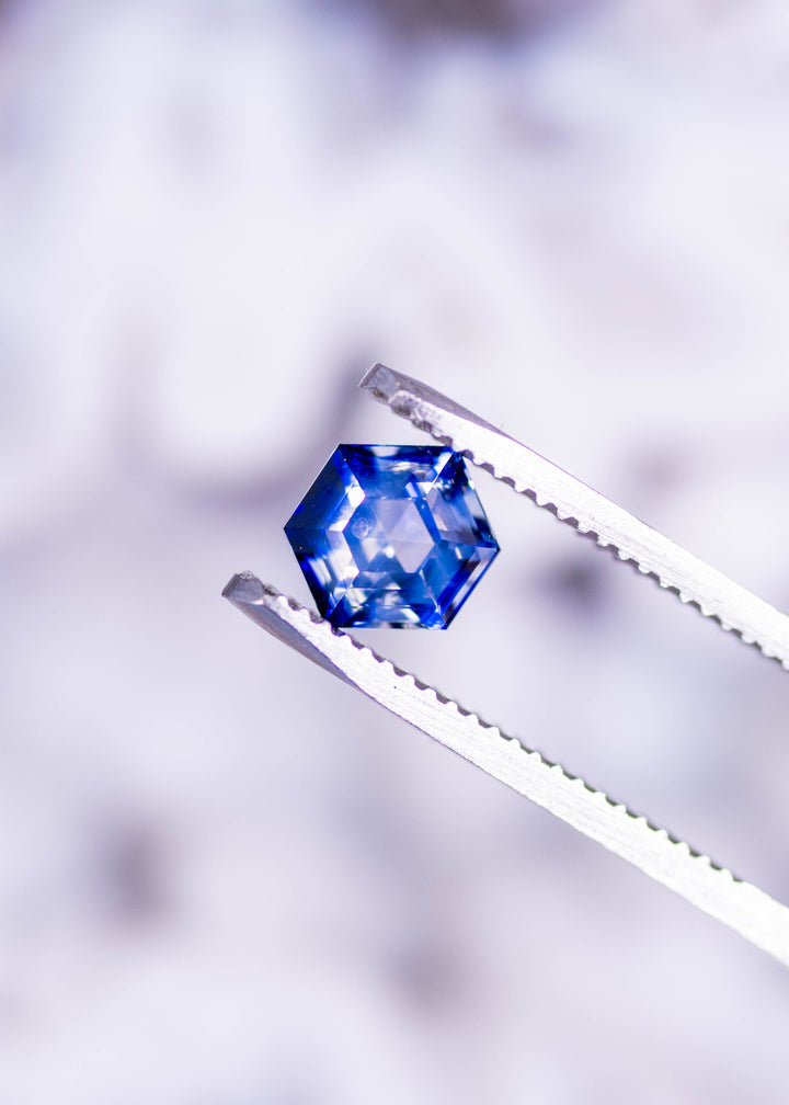 Loose blue and colorless bi‑color sapphire from Ratnapura, Sri Lanka held in jeweler’s tweezers, displaying the custom geometric hexagon step cut and crisp facet pattern.