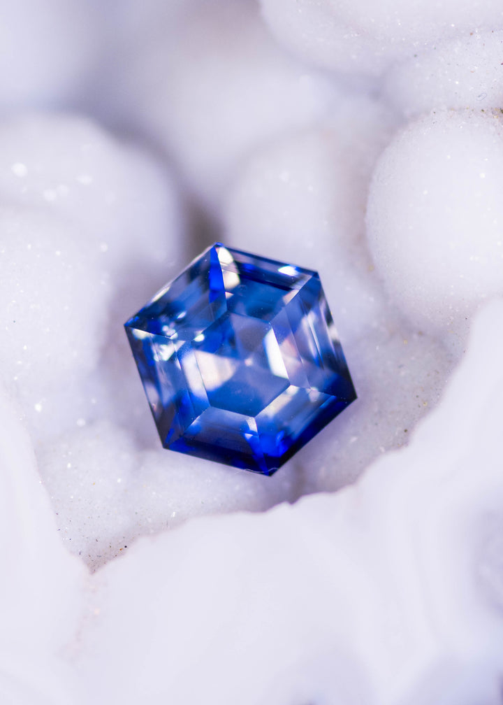Loose bi‑color blue and colorless sapphire from Ratnapura, Sri Lanka shown face‑up resting on a white crystal surface, highlighting the custom geometric hexagon step cut and natural color zoning.