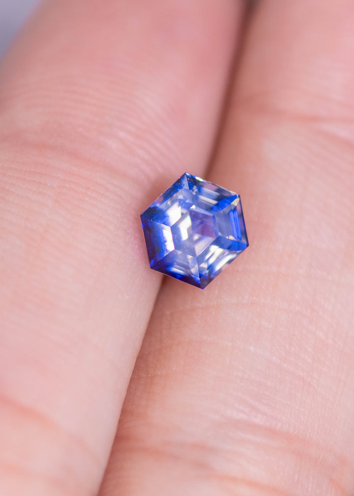 Loose bi‑color blue and colorless sapphire resting on a fingertip for scale, showing the custom geometric hexagon step cut and natural untreated color from Sri Lanka.