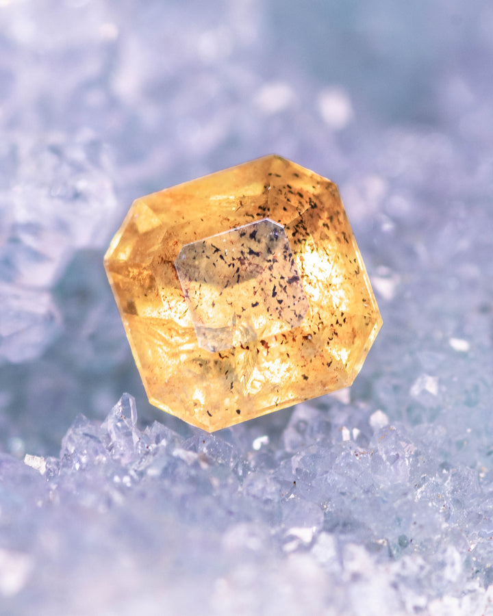 1.71 carat pepper yellow orange sapphire resting on icy blue crystals, showing natural inclusions and warm golden tones in a precision Asscher cut, untreated gemstone from Umba River Valley, Tanga, Tanzania.