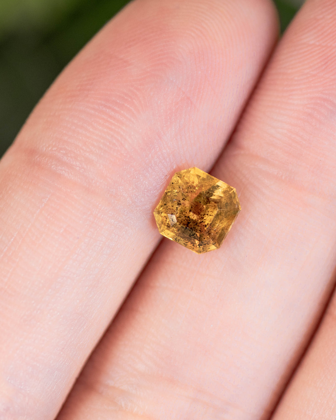 Close-up of 1.71 carat pepper yellow orange sapphire resting on a fingertip, displaying light reflection and natural inclusions within its Asscher cut, untreated gemstone from Umba River Valley, Tanga, Tanzania.