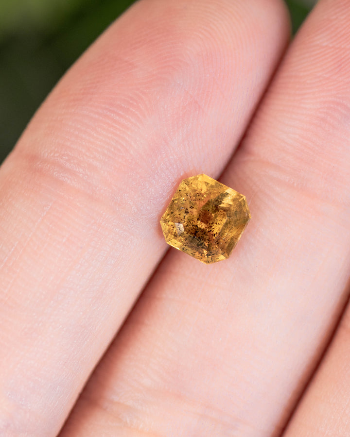 Close-up of 1.71 carat pepper yellow orange sapphire resting on a fingertip, displaying light reflection and natural inclusions within its Asscher cut, untreated gemstone from Umba River Valley, Tanga, Tanzania.