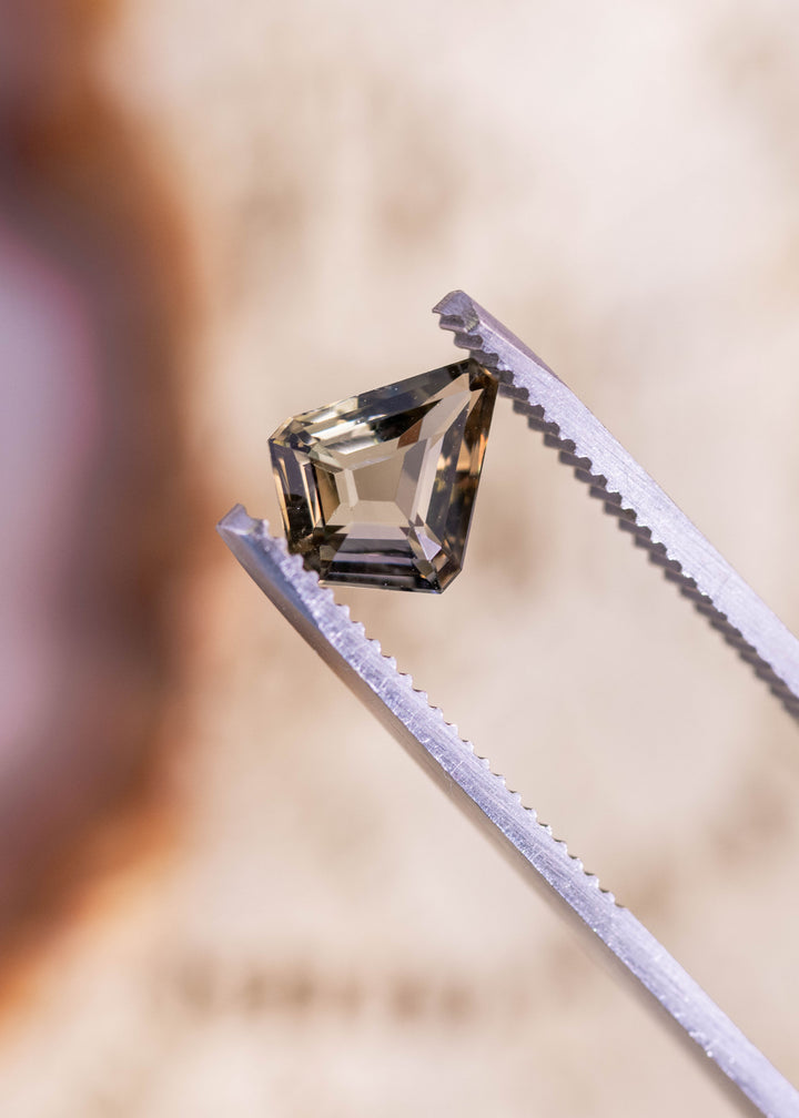 1.72 carat custom kite step cut Tanzanite in golden teal yellow colors held in gem tweezers showing geometric facets and brilliance