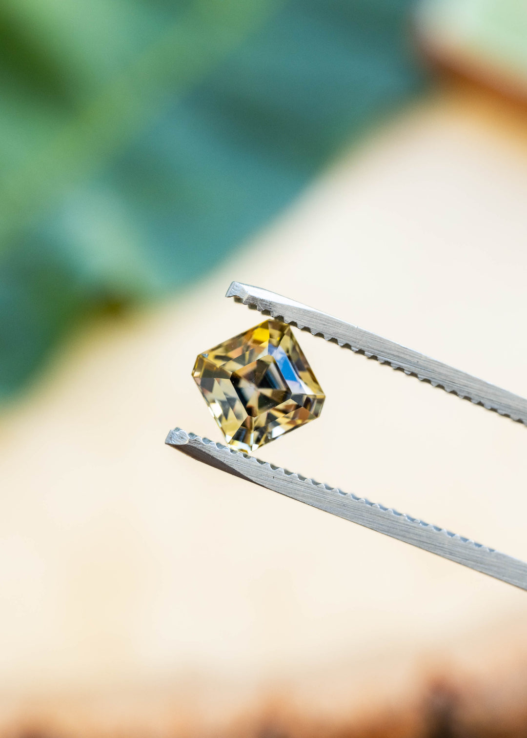 A square yellow gemstone held in silver metal tweezers against a softly blurred background with green and beige tones.