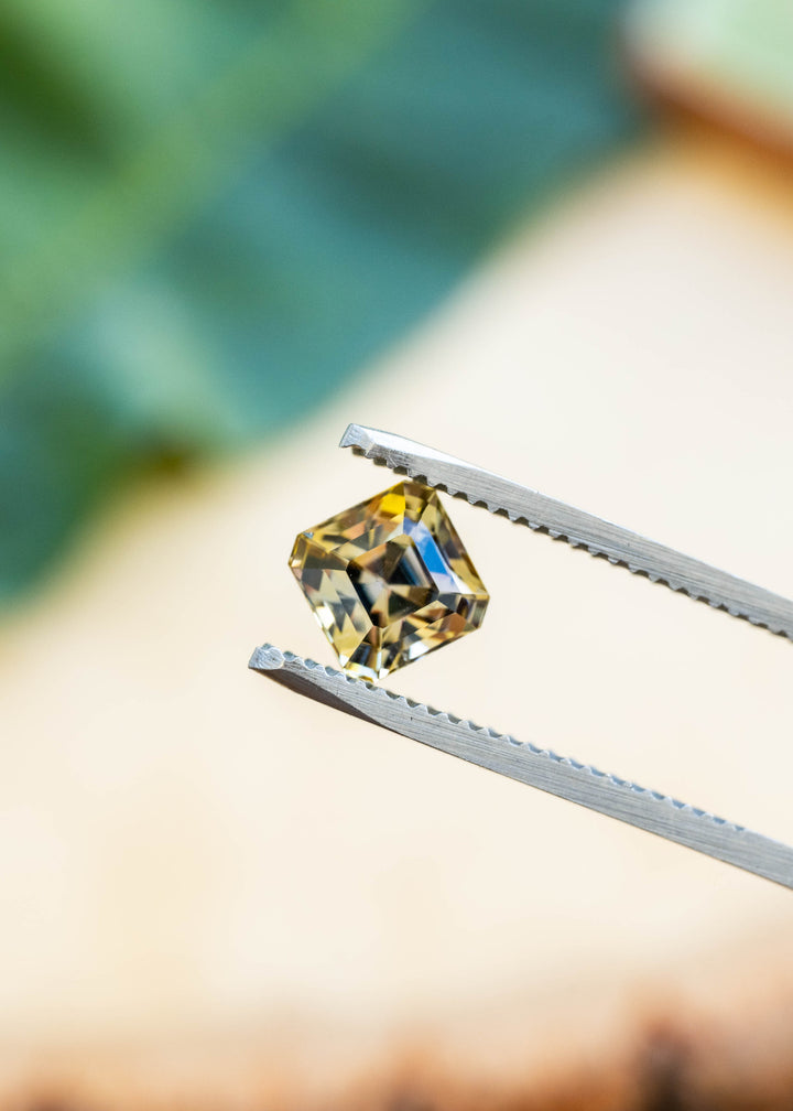 A square yellow gemstone held in silver metal tweezers against a softly blurred background with green and beige tones.
