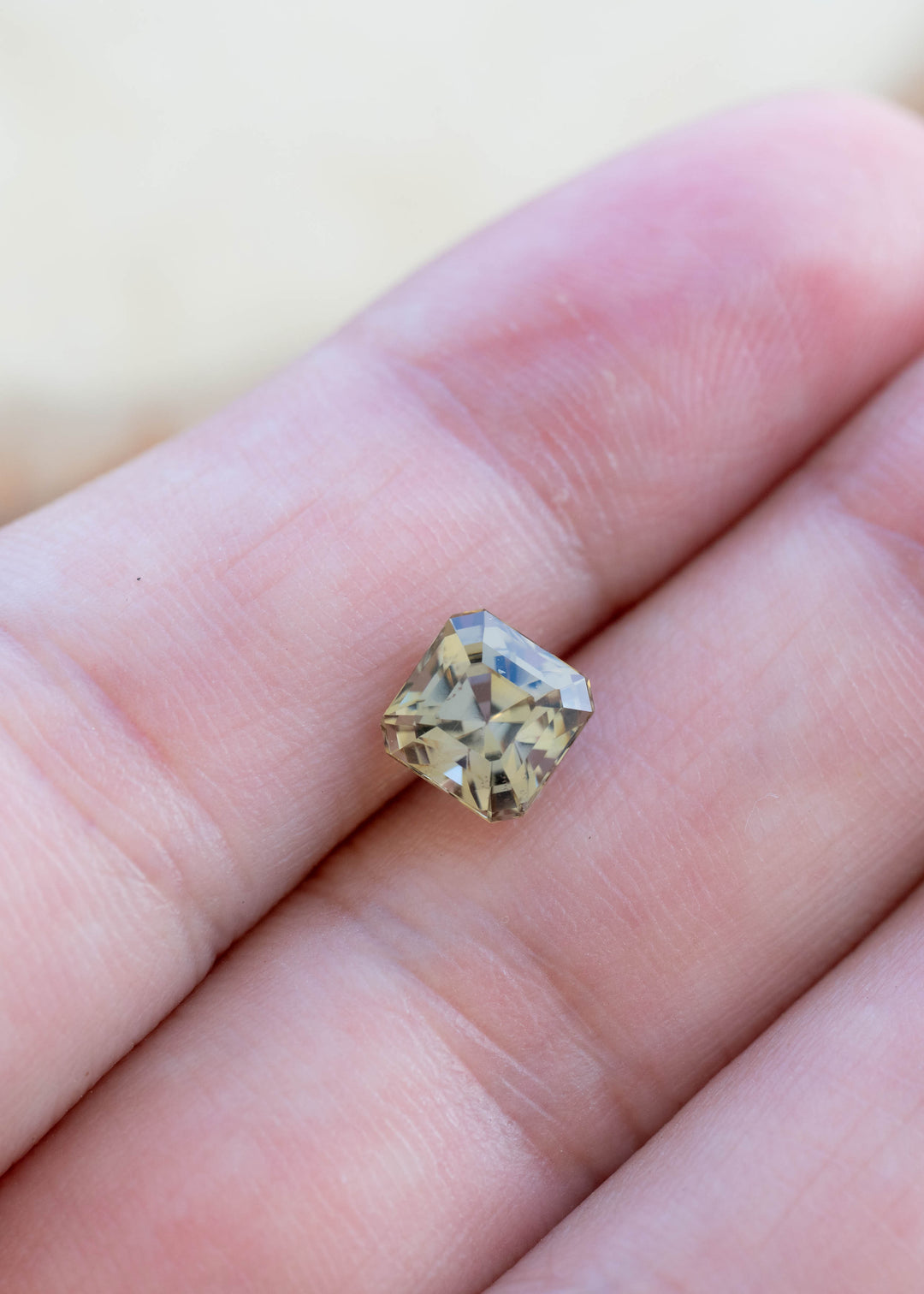 A small square-cut yellow gemstone with a halo of dust or glitter, resting on a person's fingers against a soft beige background.