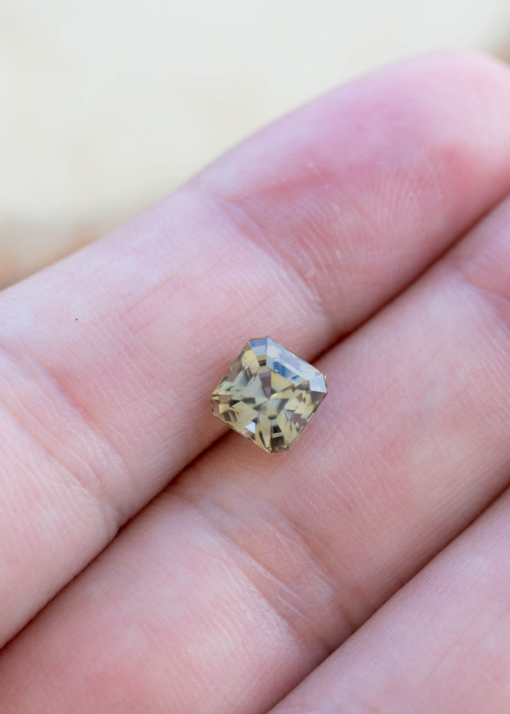 A small square-cut yellow gemstone with a halo of dust or glitter, resting on a person's fingers against a soft beige background.