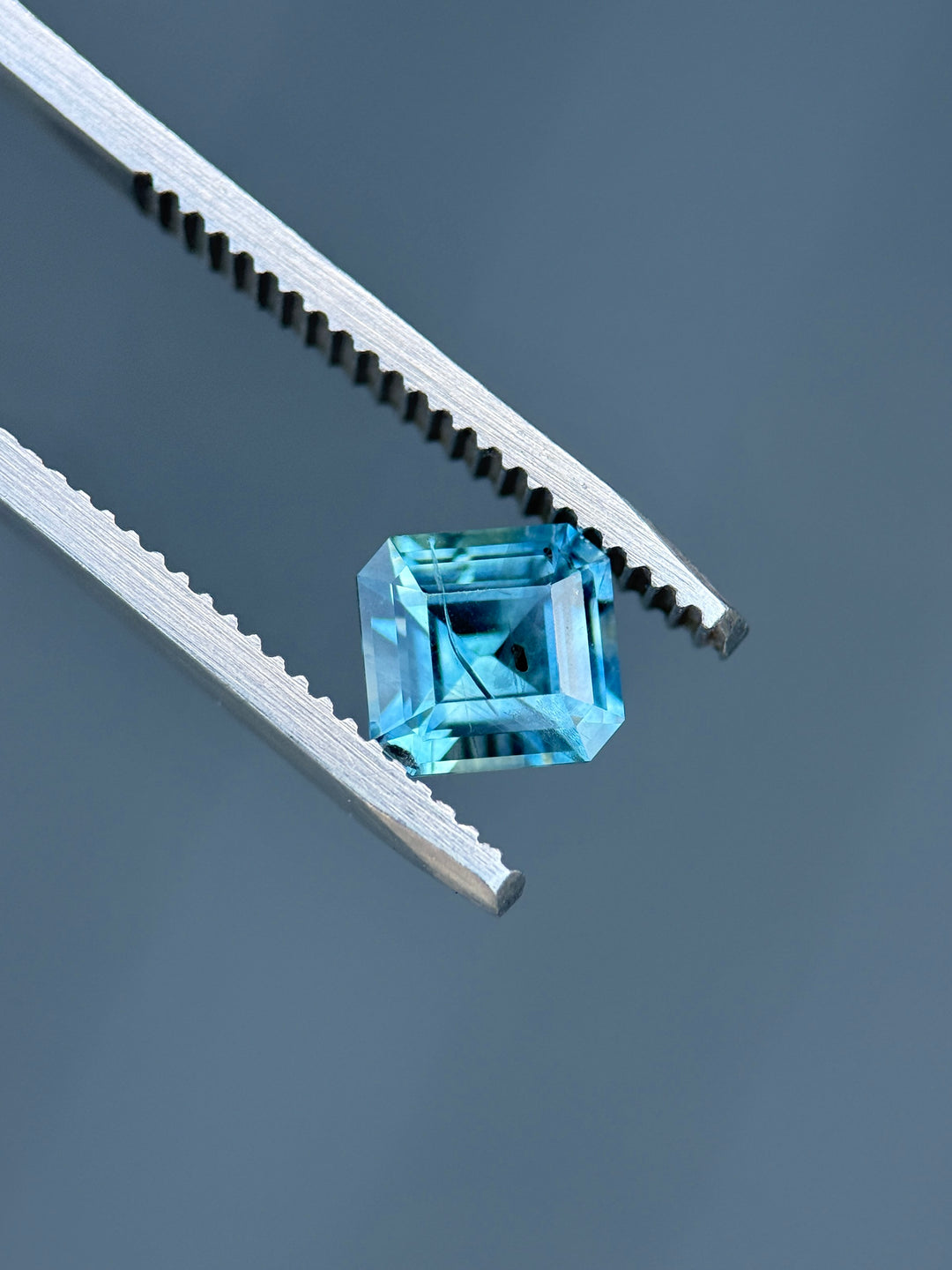 Square Asscher cut Montana sapphire held in tweezers, highlighting geometric faceting and teal-blue color.