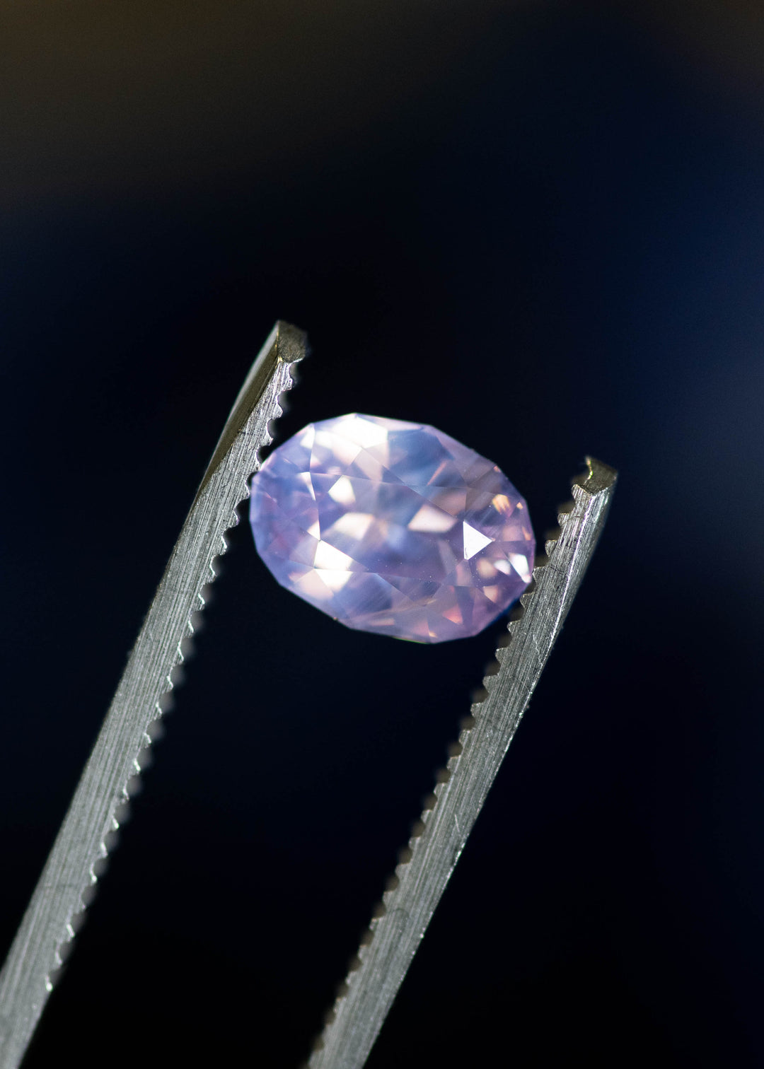 Loose opalescent pink sapphire held in tweezers, showing crisp oval facet symmetry and subtle violet undertones from Sri Lanka.