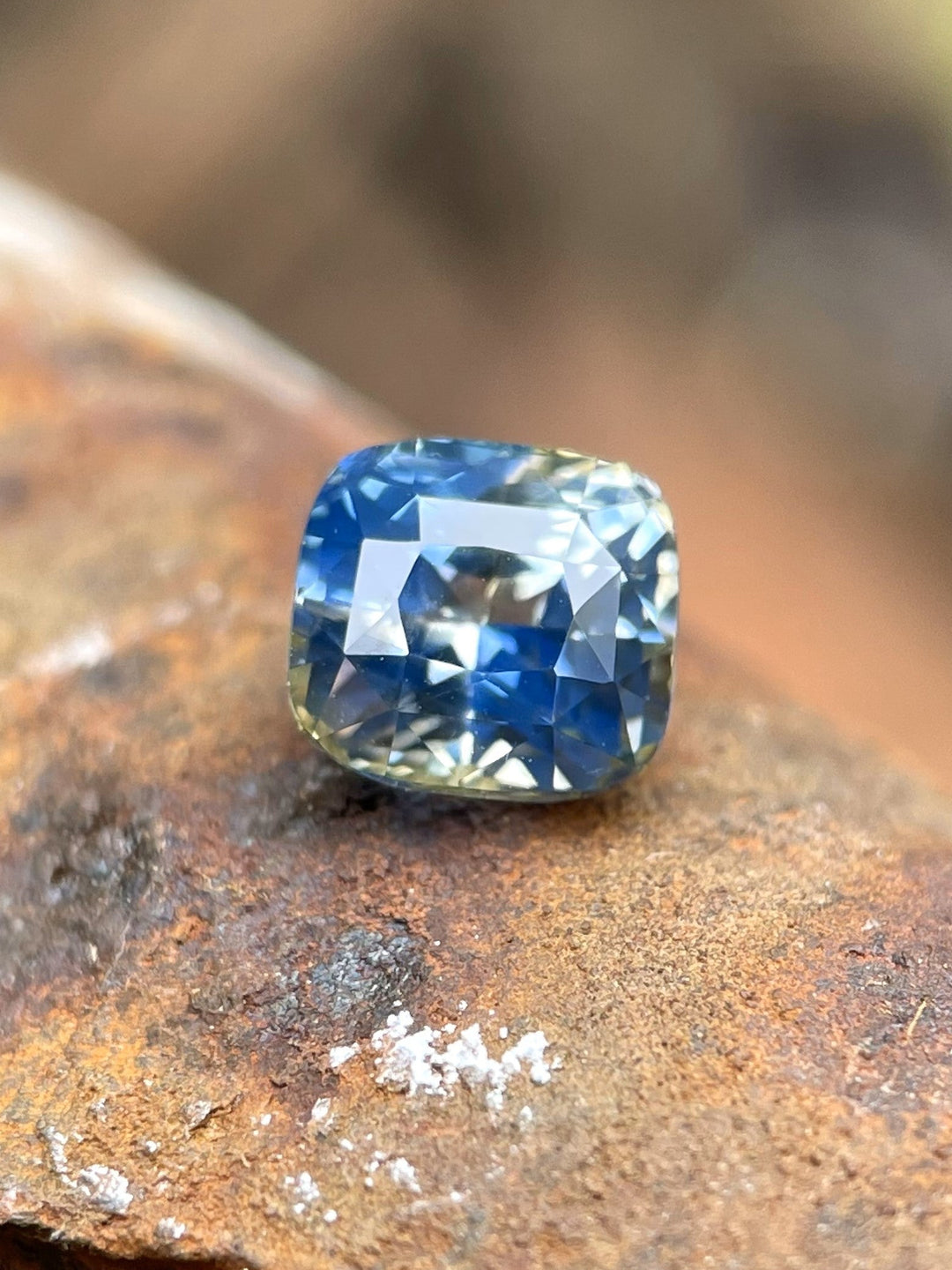 Close-up of a 1.78-carat Madagascar Parti Sapphire displaying a captivating mix of yellow, colorless, denim, and Ceylon blue hues. The modified brilliant cushion cut highlights the gemstone's intricate color play, resting elegantly on a rustic surface.