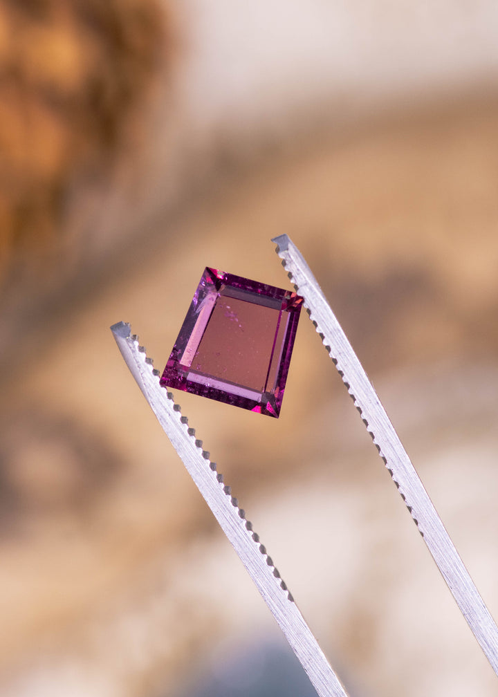 Natural pink and purple portrait cut garnet held in gem tweezers showing precision faceting and vibrant color. Perfect for bespoke jewelry bezel set pendant.