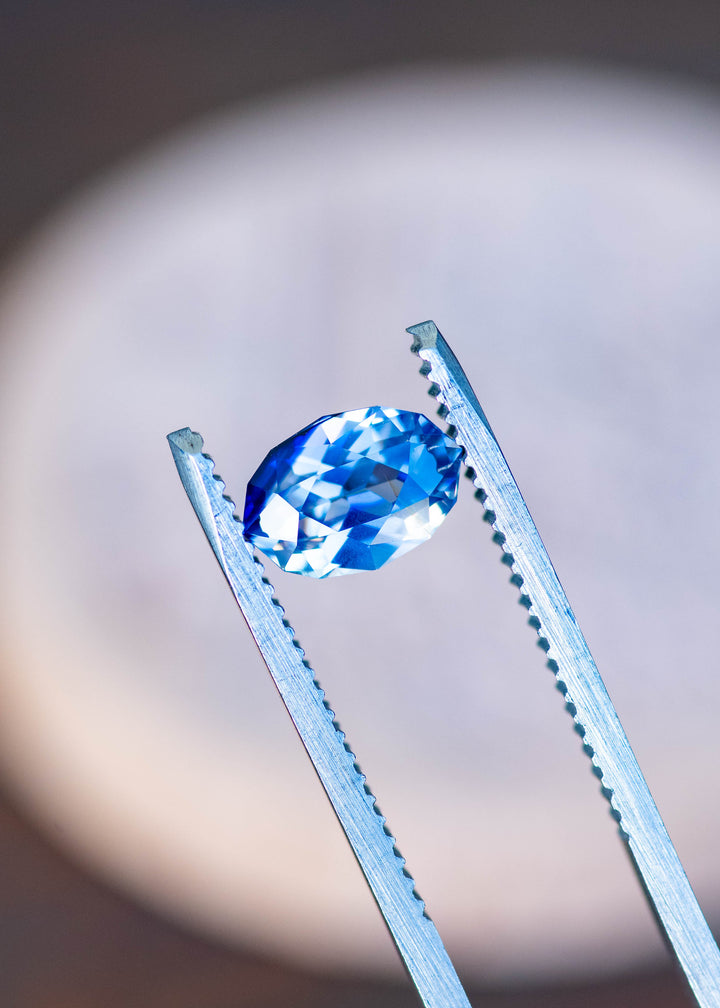 1.85ct blue and white parti sapphire in a custom moval brilliant cut from Sri Lanka held in gem tweezers under natural light.