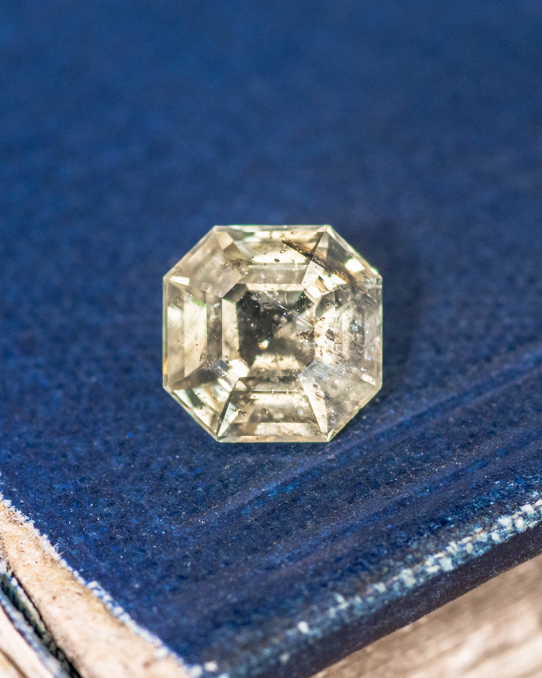 1.88 carat teal yellow sapphire displayed on a blue textured surface, showcasing Asscher cut facets and natural brilliance of this untreated gemstone from Umba River Valley, Tanga, Tanzania.