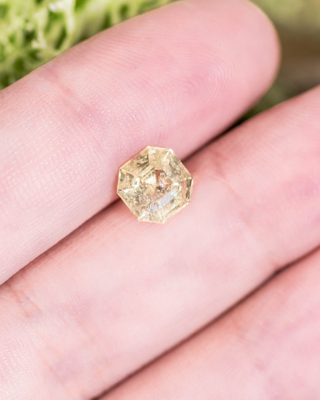 Close-up of 1.88 carat teal yellow sapphire resting on fingertip, highlighting geometric facets and natural light reflections in its Asscher cut, untreated gemstone from Umba River Valley, Tanga, Tanzania.