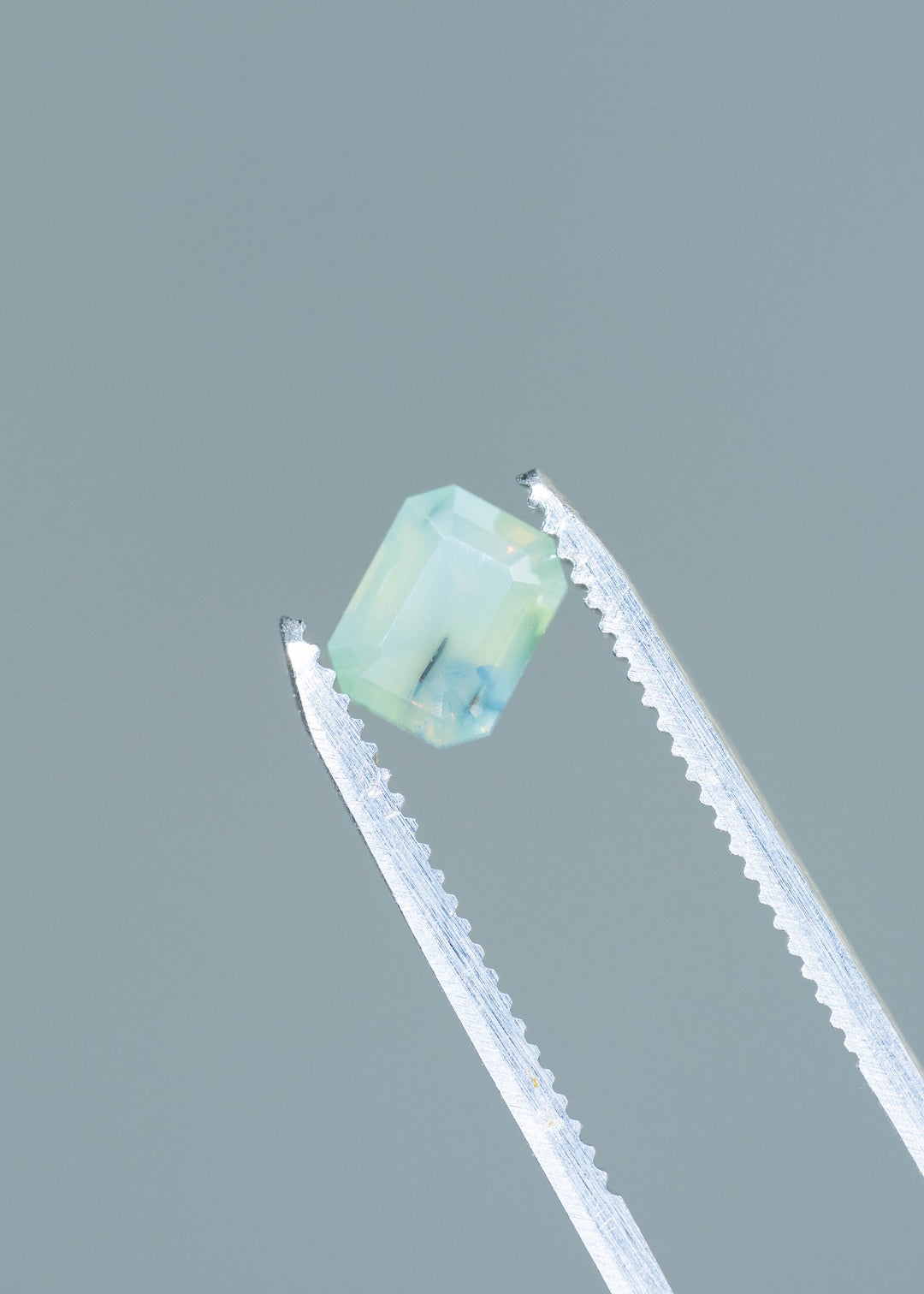 Pale seafoam-blue sapphire held at an angle between silver tweezers against a gray background. Its rectangular shape has clipped corners and broad facets, with a dark natural inclusion visible through the softly translucent center.