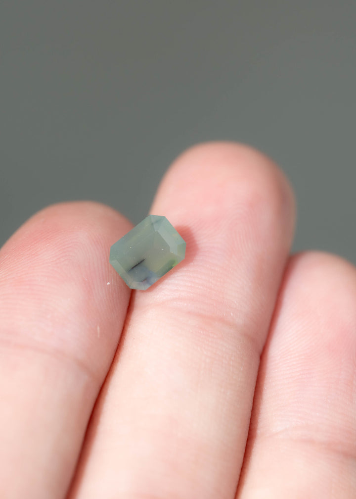 Small, pale greenish-blue emerald cut sapphire resting between fingertips. Its clipped corners and broad facets frame a softly clouded interior with a narrow dark inclusion near the lower edge.