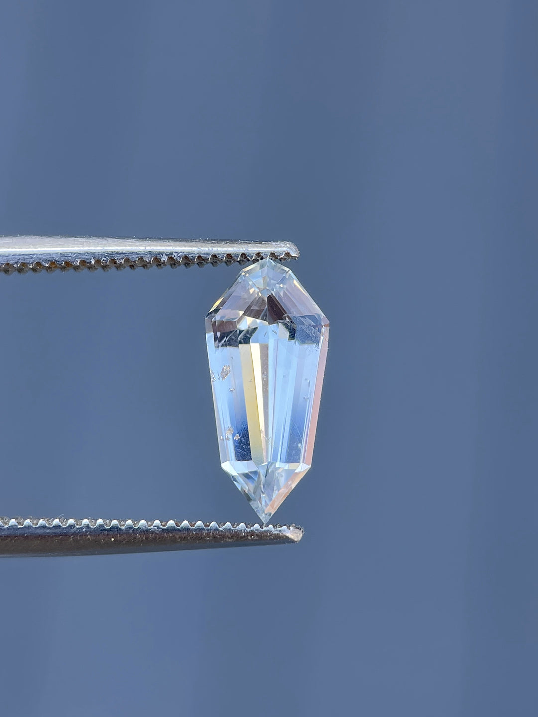 A 1.90-carat pale seafoam sapphire in a custom step cut shield shape, held in tweezers, showcasing its brilliance and elongated geometric facets.