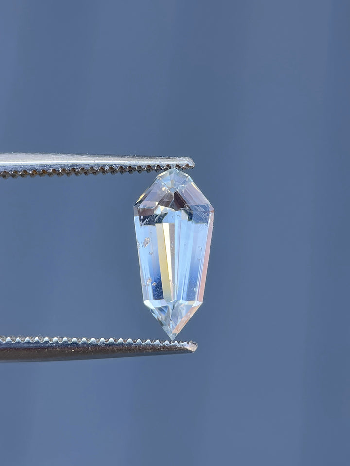 A 1.90-carat pale seafoam sapphire in a custom step cut shield shape, held in tweezers, showcasing its brilliance and elongated geometric facets.