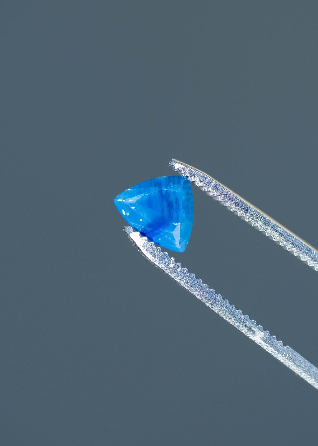 Close-up photograph of an untreated 1.91ct royal blue sapphire held in jeweler's tweezers, highlighting its custom triangular cut, opalescent appearance and visible internal texture.
