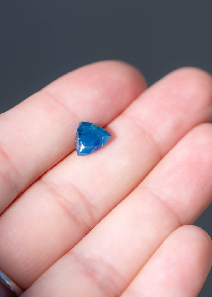 Untreated 1.91 carat royal blue sapphire from Nigeria displayed on fingers for scale. The custom trillion cut measures 8.54 × 7.13 mm and features distinctive opalescent blue coloration.