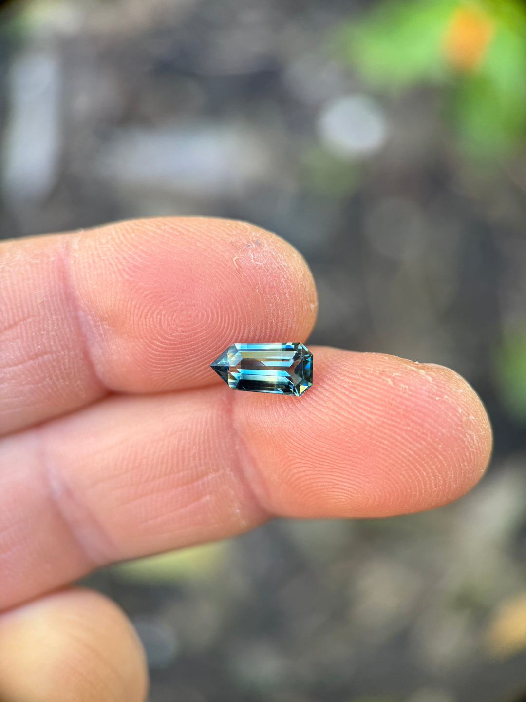 Loose yellow blue green parti sapphire from Diego Madagascar shown on a fingertip highlighting the elongated shield shape and step cut facet structure.