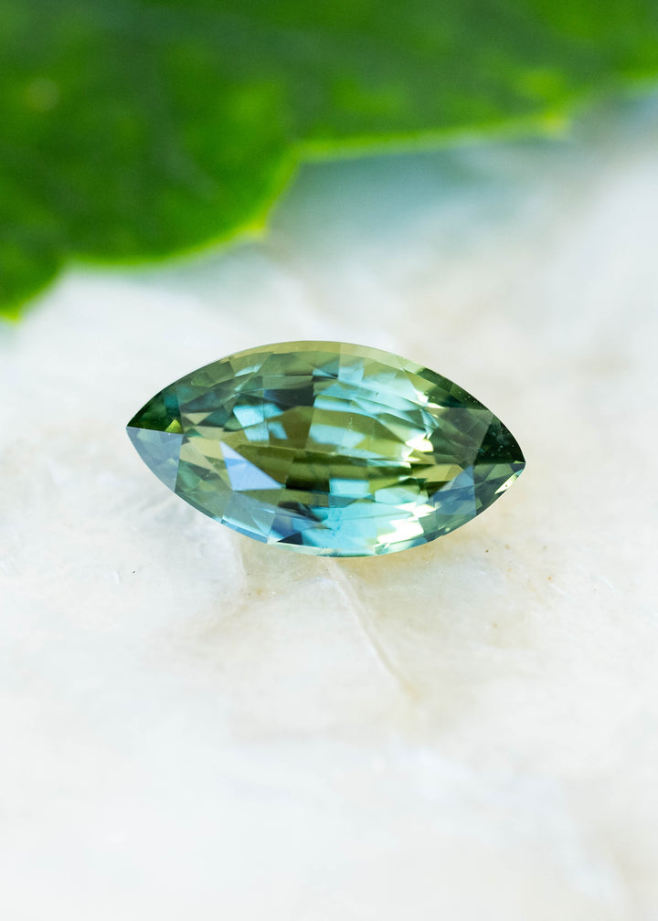 Natural 2.03 carat teal blue yellow parti sapphire from Diego, Madagascar, photographed on a pale surface beneath a green leaf. Its custom octagonal radiant mix cut displays turquoise blue, olive green, and golden-yellow reflections.