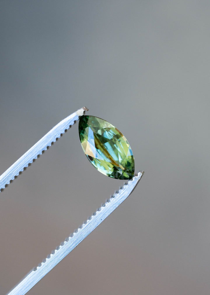 Close-up of a 2.03ct heated parti sapphire from Madagascar held in jeweler's tweezers. Its multicolored faceting reveals bright teal-blue reflections, olive-green zoning and golden-yellow accents.