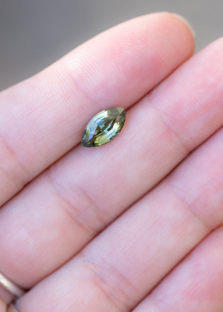 Natural 2.03 carat teal blue yellow parti sapphire from Diego, Madagascar, photographed on fingers for scale. The gemstone displays an elongated, pointed silhouette with contrasting green, blue and yellow facets.
